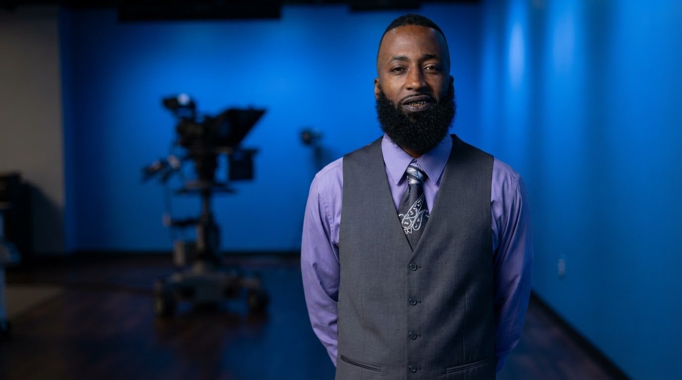 Delano standing front-facing three-quarter, hands behind his back, in the grey vest, lavender shirt and patterned tie