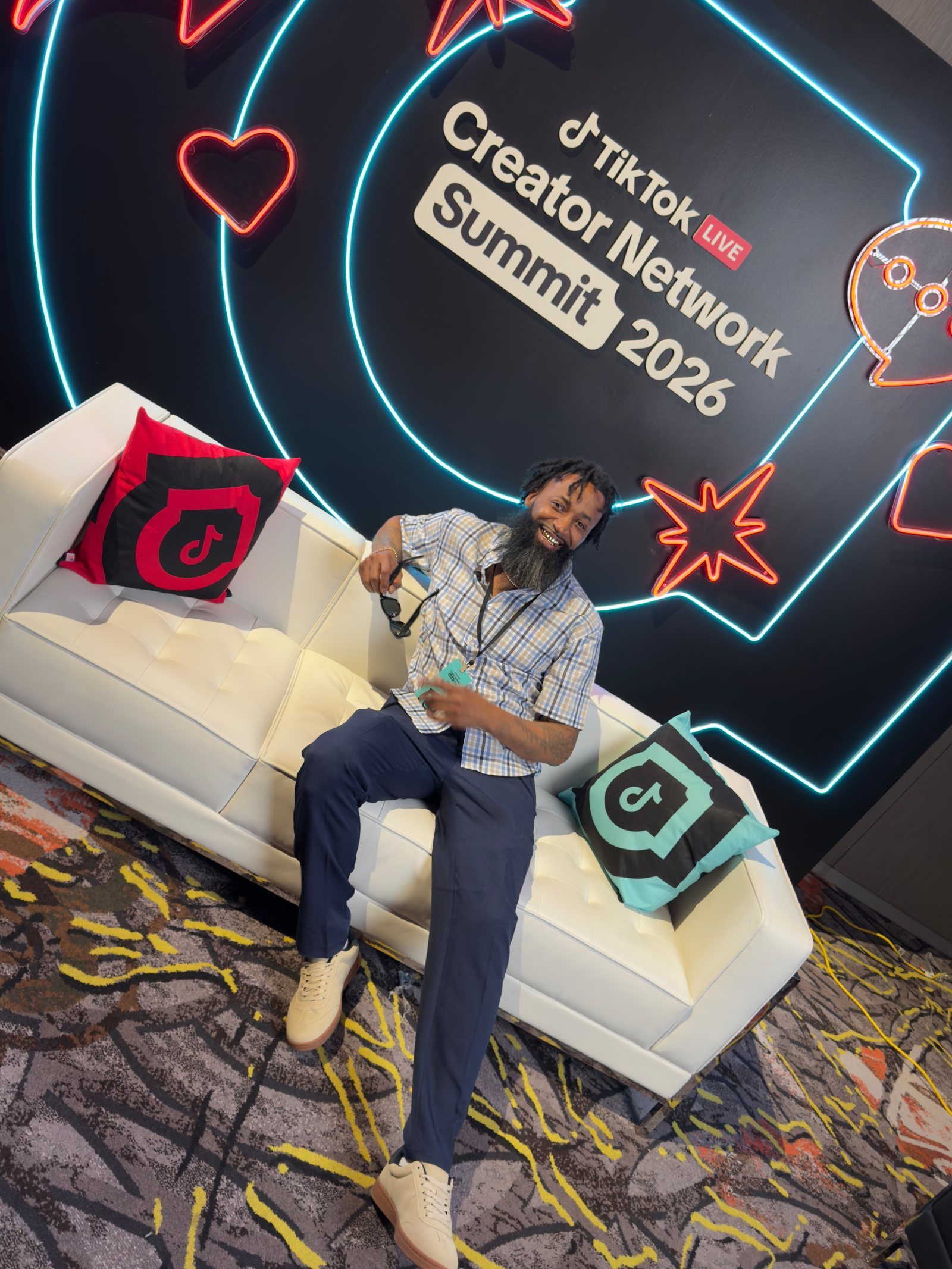 Delano seated on the white tufted couch at the creator-network summit with the branded pillows and neon signage behind him